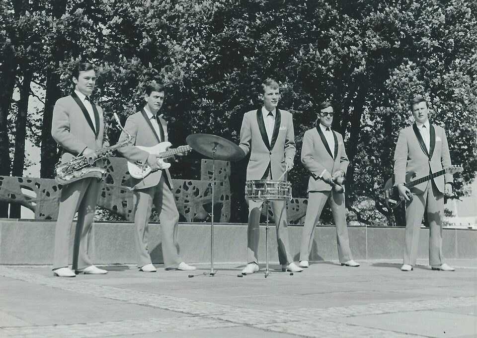 Schwarzweißfoto: fünf Männer in hellen Anzügen mit dunklem Revers stehen auf einem Platz, mit Saxofon, zwei Gitarren und Schlagzeug, dahinter Bäume.