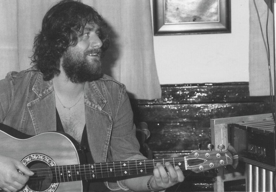 Schwarzweißfoto: ein bärtiger Mann mit lockigem Haar in Jeansjacke spielt eine Akustikgitarre und blickt zur Seite, rechts ein Gerät mit Metallstäben.