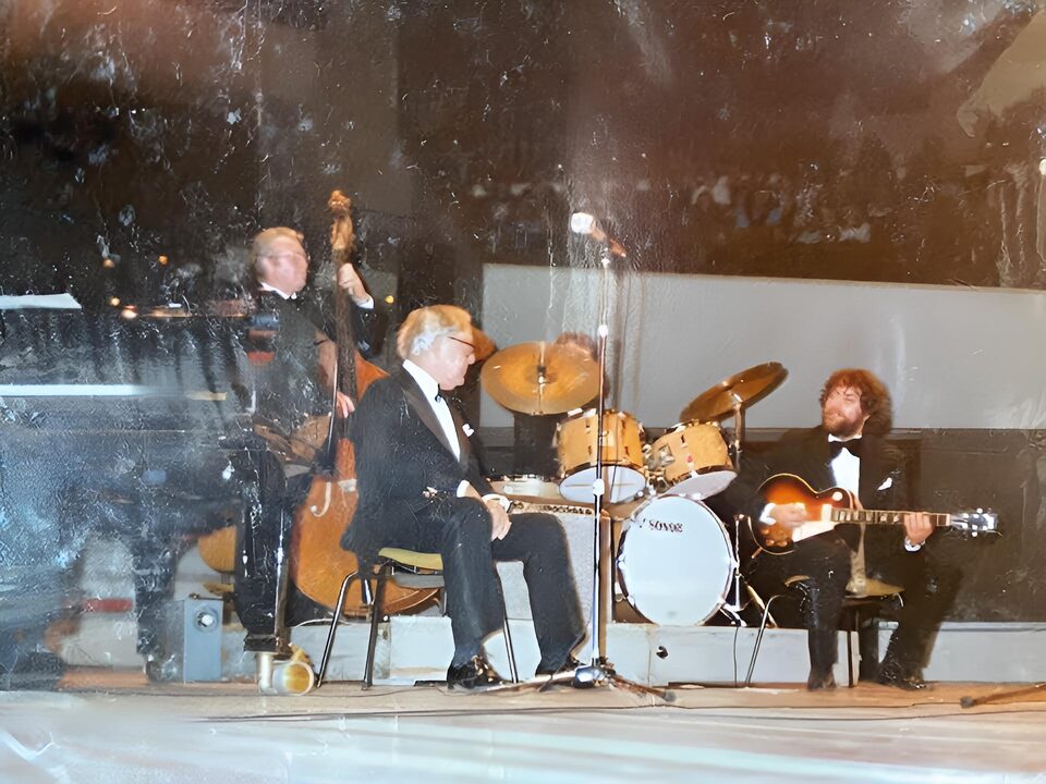 Stark beschädigter Farbabzug einer Bühnenszene: ein Bassist, ein sitzender Mann im Smoking, ein Schlagzeug und rechts ein Gitarrist im Scheinwerferlicht.