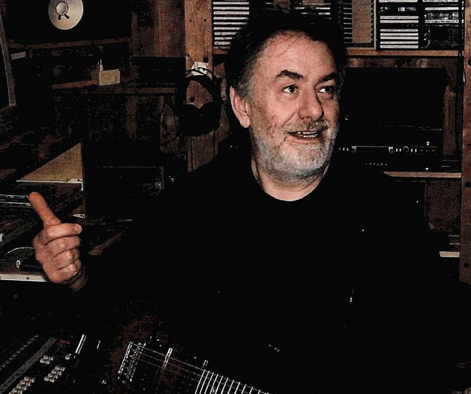 Farbfoto: Harry Pepl mit grauem Bart und dunklem Oberteil sitzt in seinem Tonstudio zwischen Holzregalen, Kopfhörern und Studiogeräten, hebt den Daumen und blickt zur Seite; unten im Bild der Hals einer E-Gitarre.