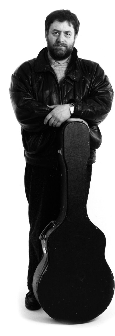 Schwarzweißes Ganzkörperporträt vor weißem Grund: Harry Pepl in schwarzer Lederjacke und dunkler Hose lehnt mit verschränkten Armen auf einem Gitarrenkoffer und blickt ernst in die Kamera.