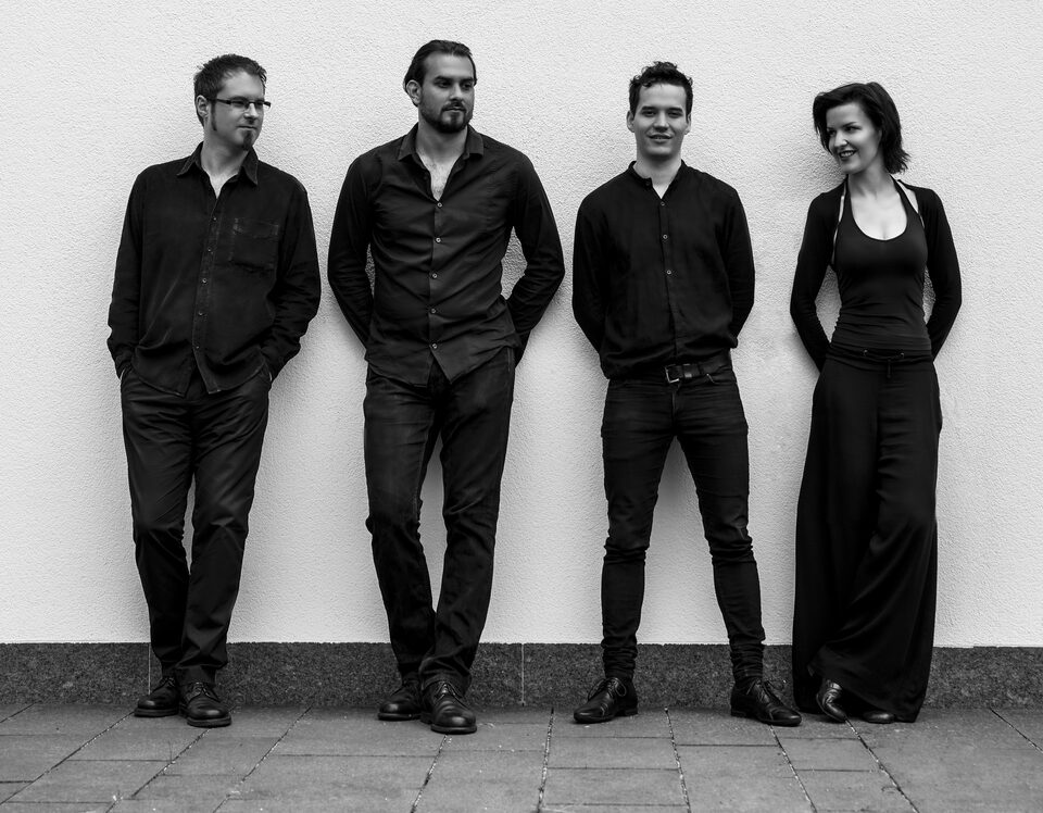 Schwarzweißes Gruppenbild: drei Musiker und eine Musikerin stehen nebeneinander vor einer weißen Wand auf Pflaster und blicken in die Kamera.